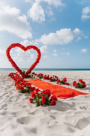 Red heart-shaped floral arch and walkway set up on a tropical beach with white sand and blue ocean views.