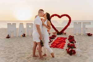 Romantic beach proposal scene with large letters and heart shape made of red roses on sandy shore at sunset.
