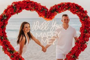 A romantic beach proposal setup with a neon 'Will You Marry Me?' sign inside a large heart made of red roses.