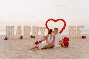Romantic beach proposal setup with illuminated 'MARRY ME' letters and red heart decoration on sand at sunset.