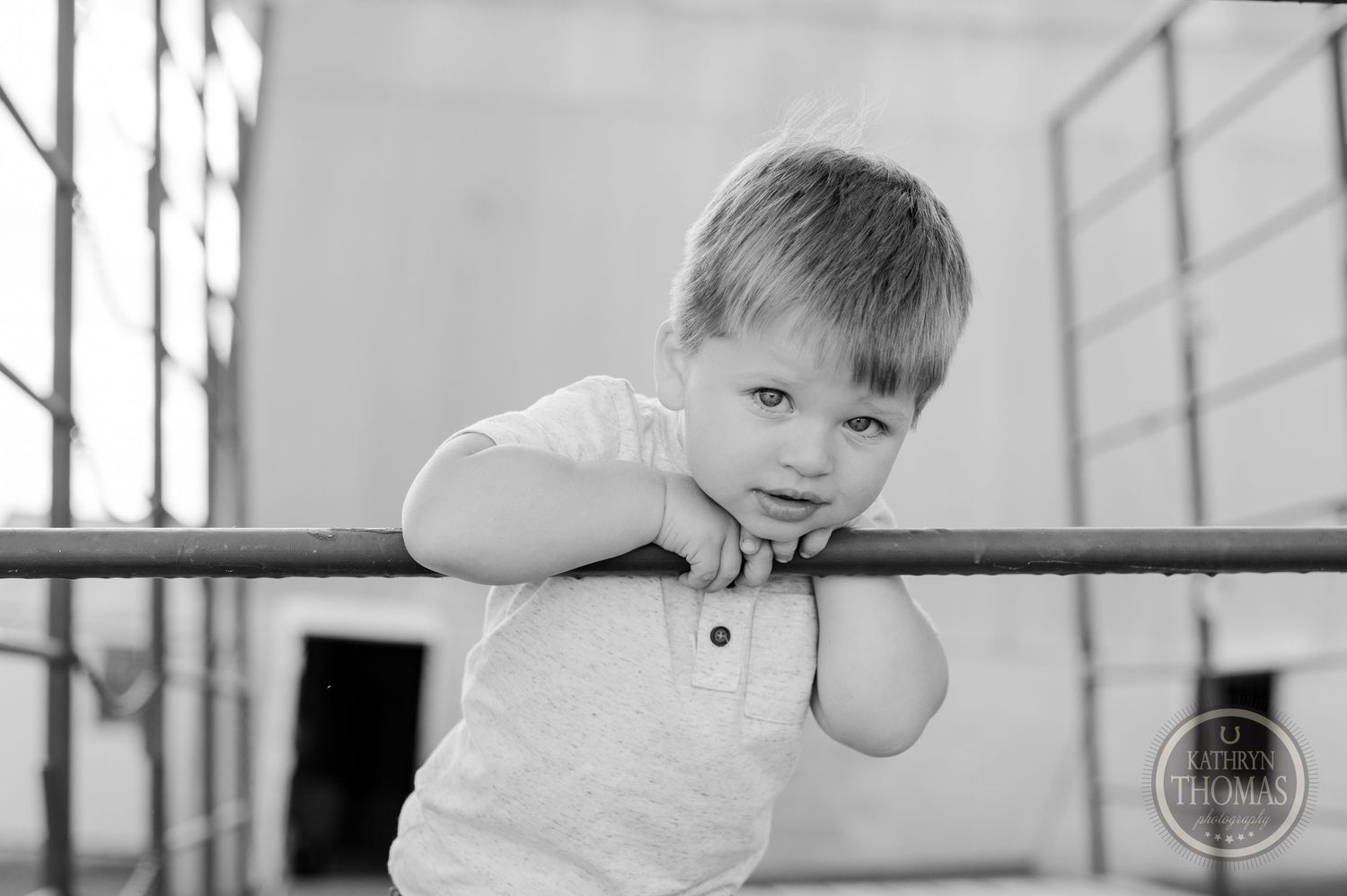 Child in white shirt leans against metal railing in black and white portrait.