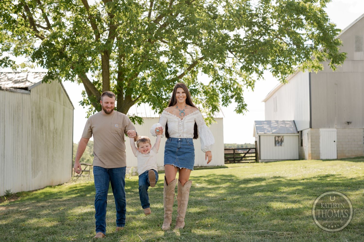 Family strolling together on grass with white barn and tree in rustic farm setting.