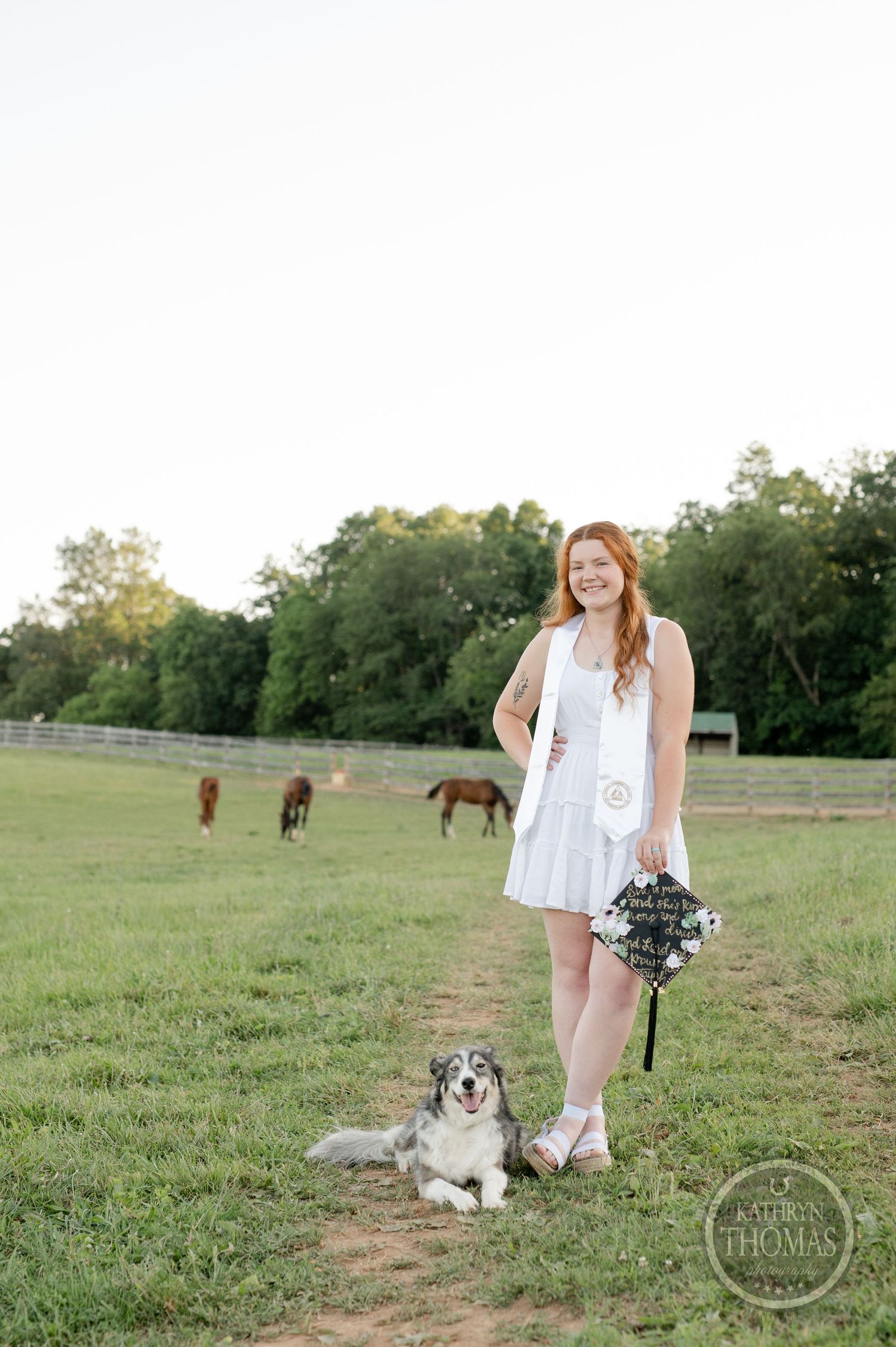 Graduate holds bouquet while standing with dog in field as horses graze in distance.