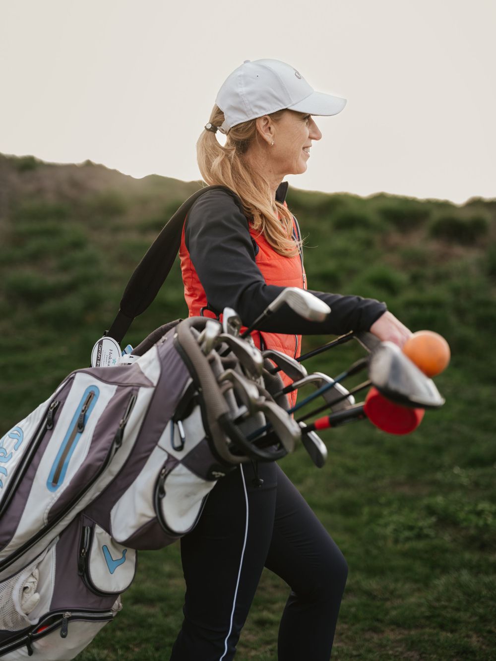 A golfer carrying a golf bag walks across a grassy course wearing orange and black athletic wear with a white cap.