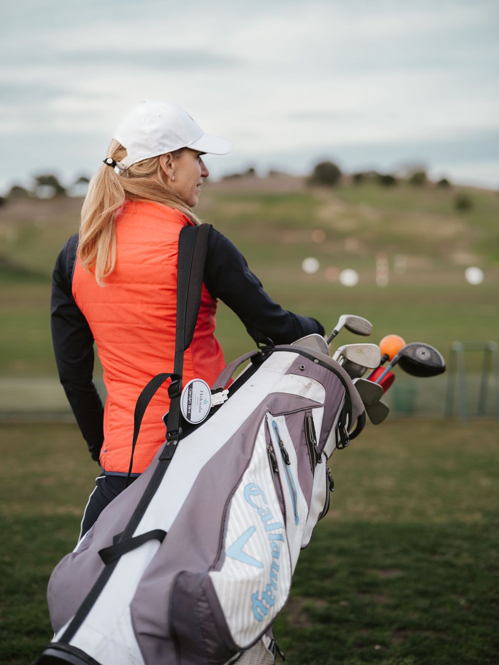 Golfer in bright orange vest and black sleeves stands with golf bag on rolling hillside course.