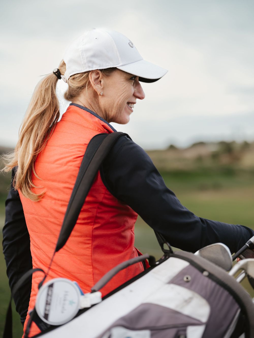 Golfer in orange vest smiles while holding golf bag against cloudy sky.