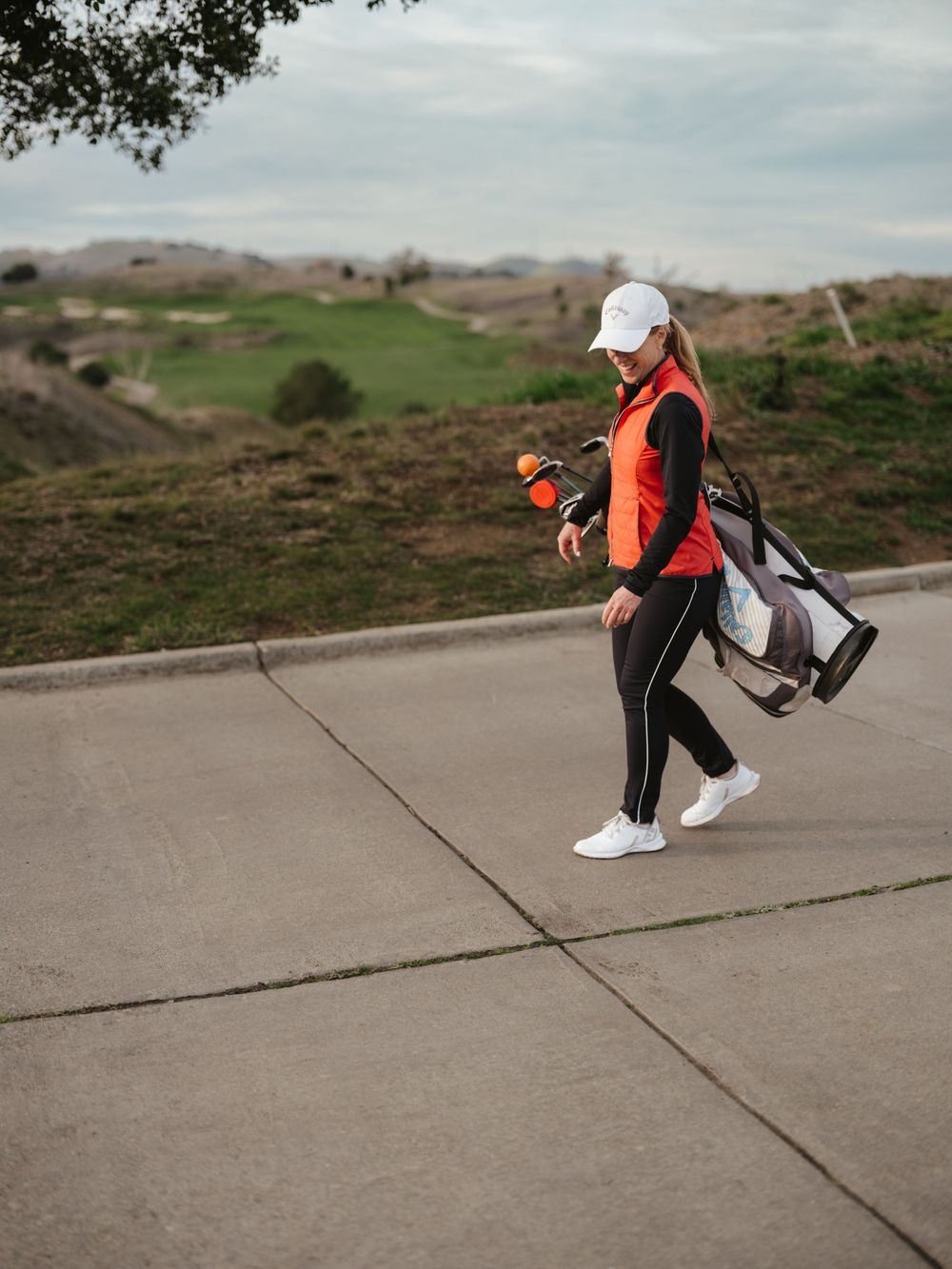 Golfer walking along concrete path carrying golf bag with rolling hills in background.