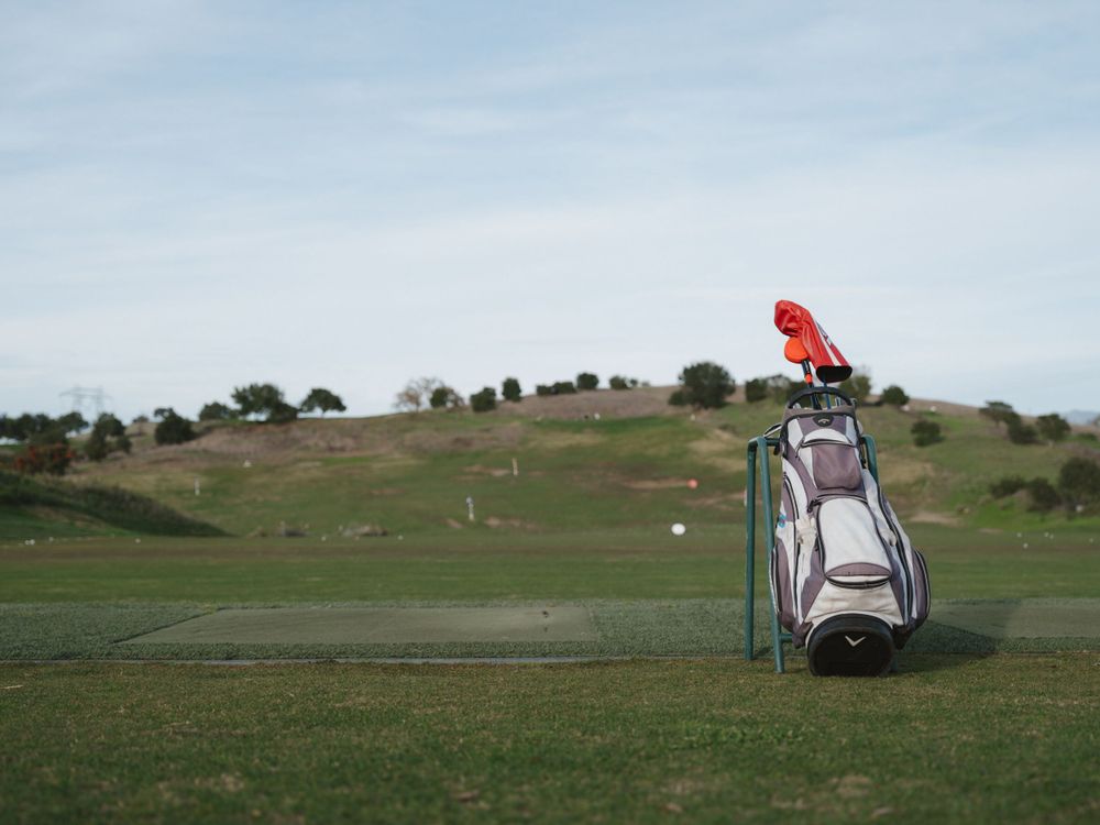 A golf bag with clubs stands on a green golf course under blue skies with hills in the background.