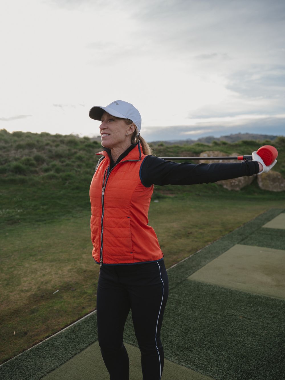 A golfer in an orange vest stretches their arms out on a golf course practice area.