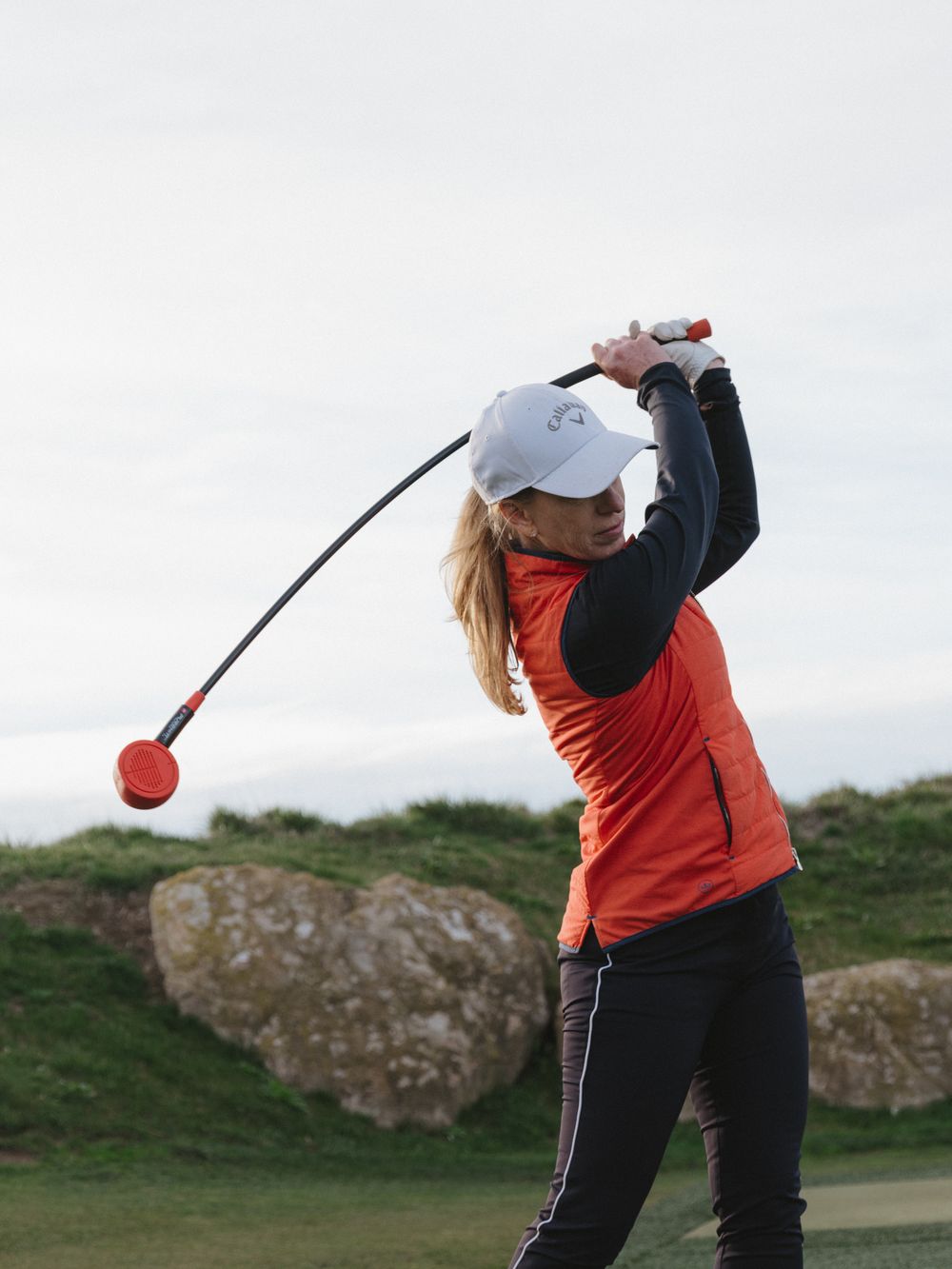 A golfer in orange vest swings a training club at full extension against a cloudy sky.