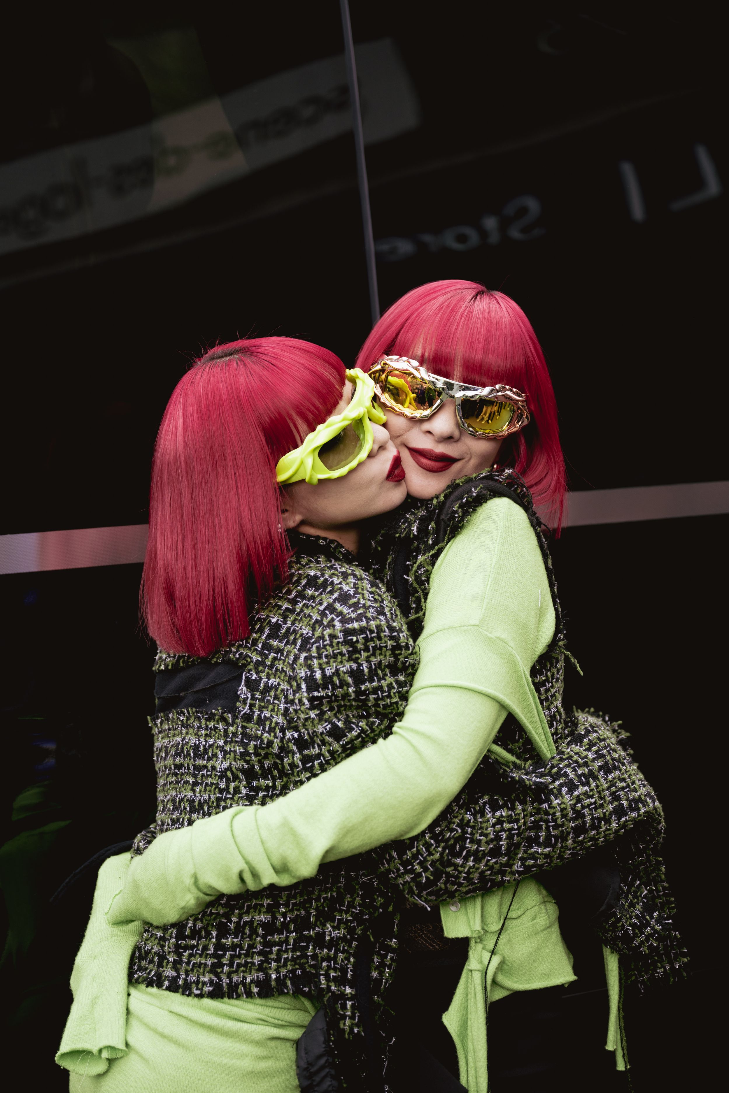 Two people with bright pink hair and neon accessories embrace while wearing matching streetwear outfits.