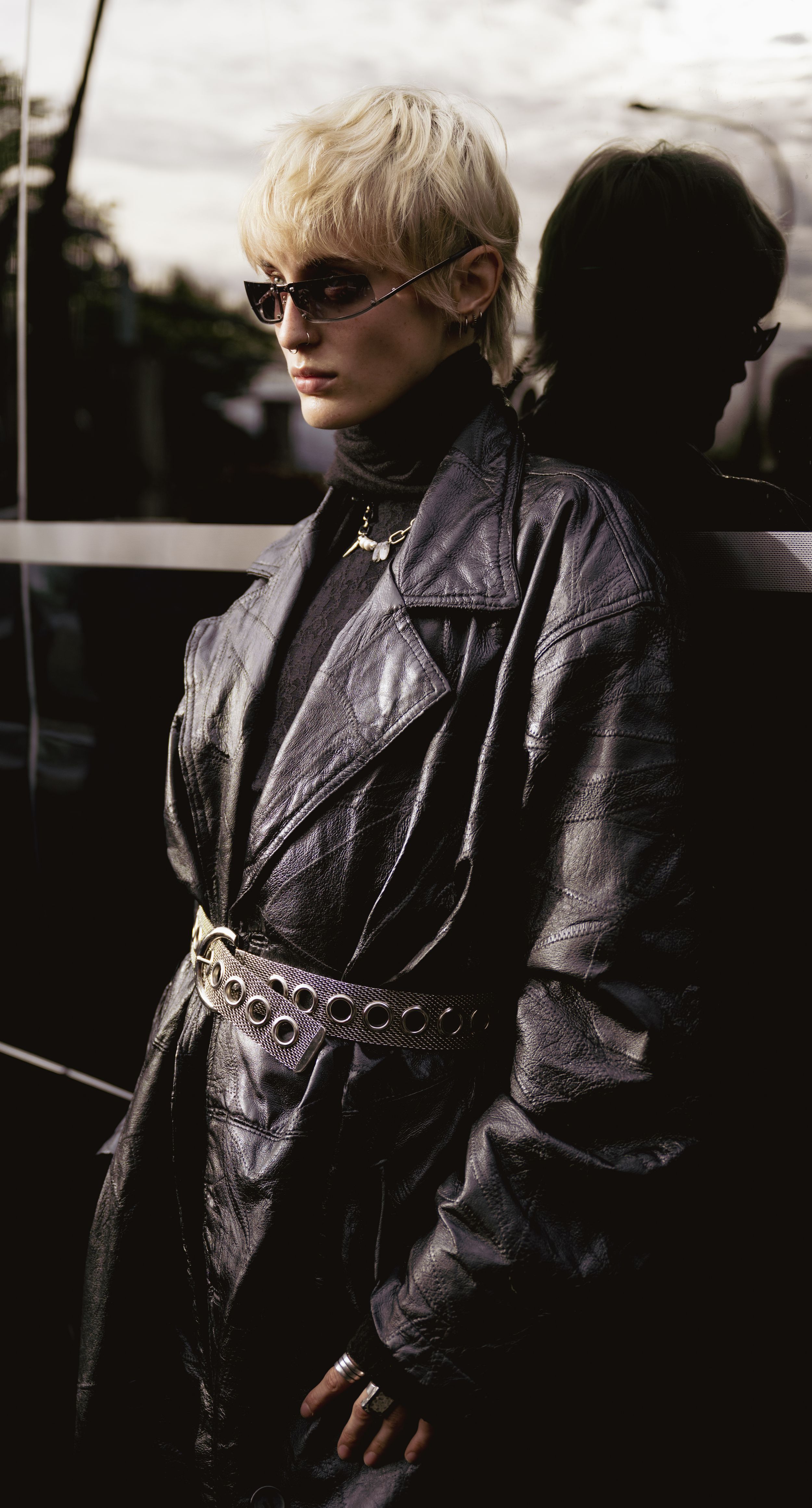 Person in edgy black leather jacket and sunglasses stands against dark background in dramatic lighting.