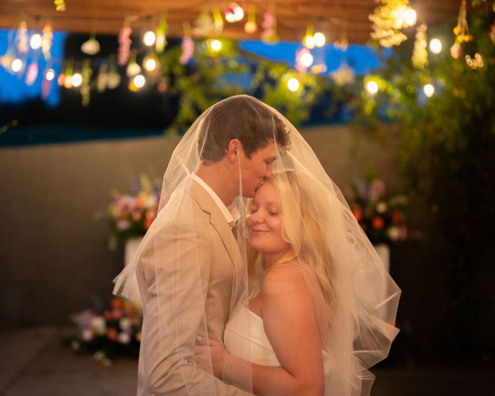 A couple embraces during their evening wedding celebration with twinkle lights and decorations glowing in the background.