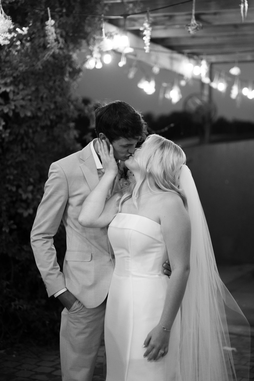 A tender black and white wedding moment captured at dusk under string lights, with the bride in a sleek gown and flowing veil.