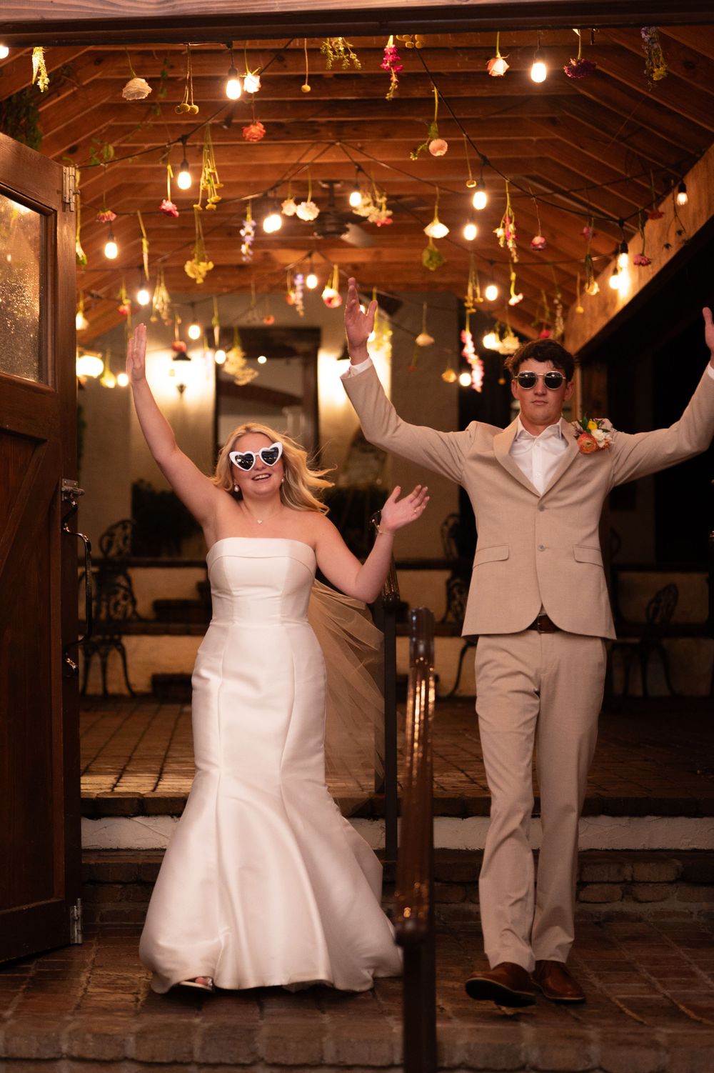Two wedding celebrants wearing sunglasses dance joyfully under hanging lights at a rustic Tennessee reception venue.