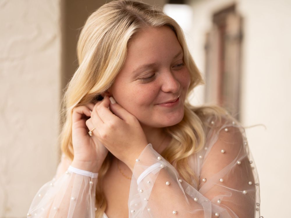 A bride in a sheer white polka dot blouse smiles while touching their blonde hair in soft lighting.