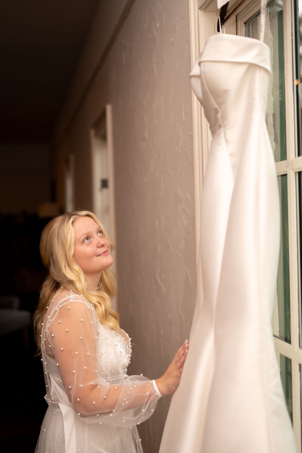 A wedding dress hangs by a window as the bride admires the elegant white gown in soft natural light.