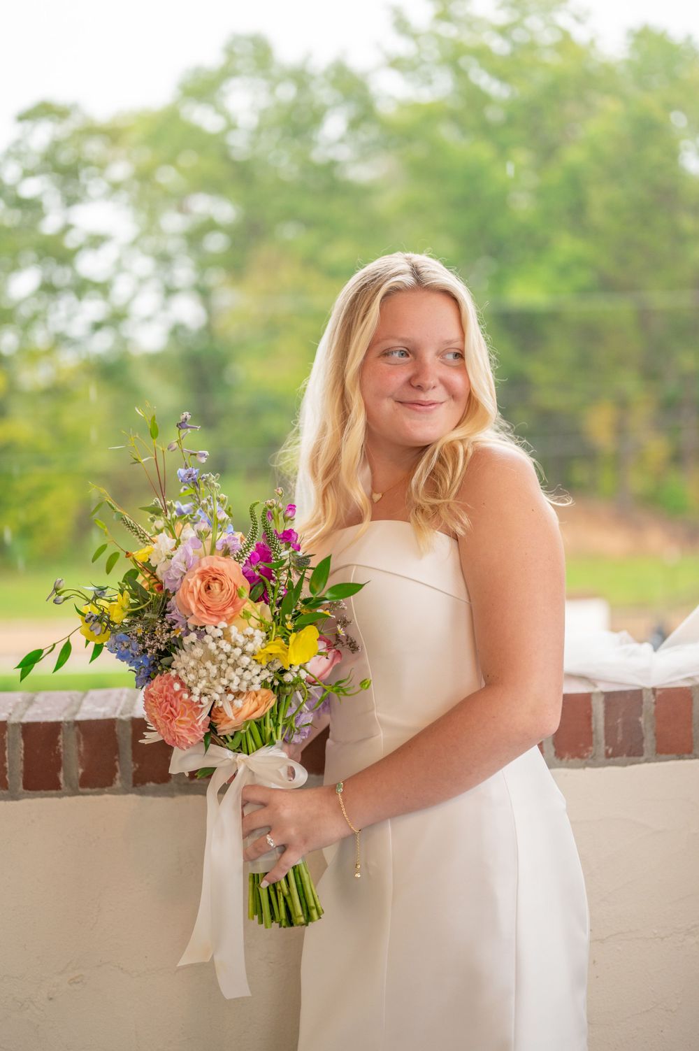 A bride in a white wedding dress holds a vibrant bouquet of mixed flowers including peach roses and purple blooms.