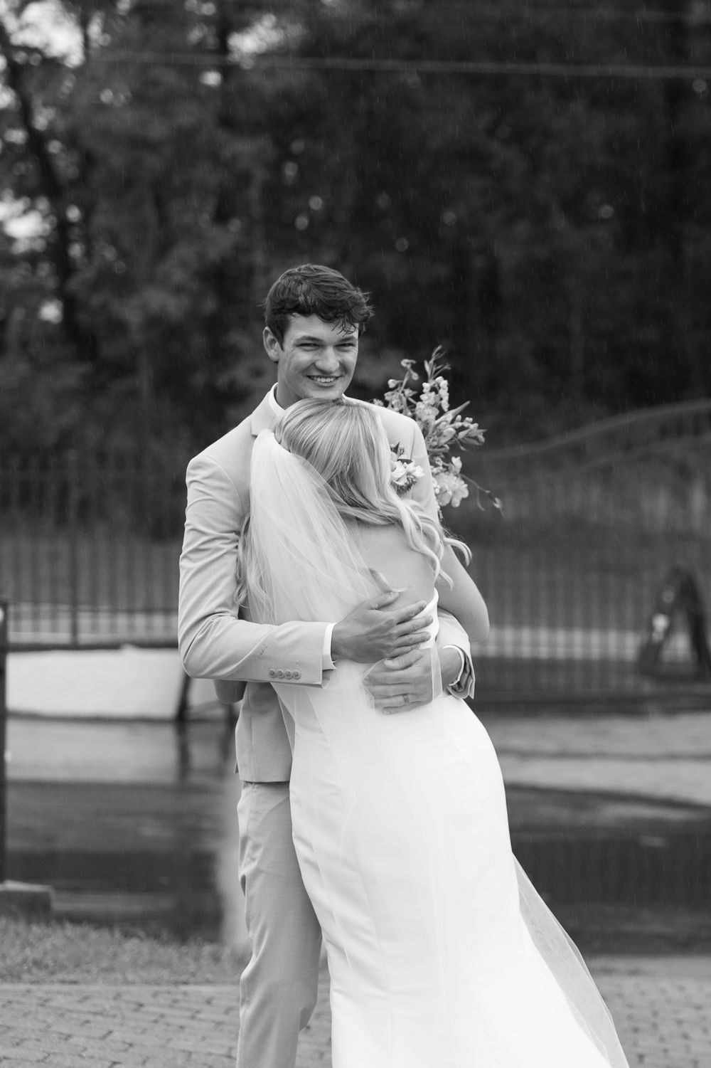 A black and white wedding photo of a couple embracing outdoors, with the bride holding a floral bouquet.