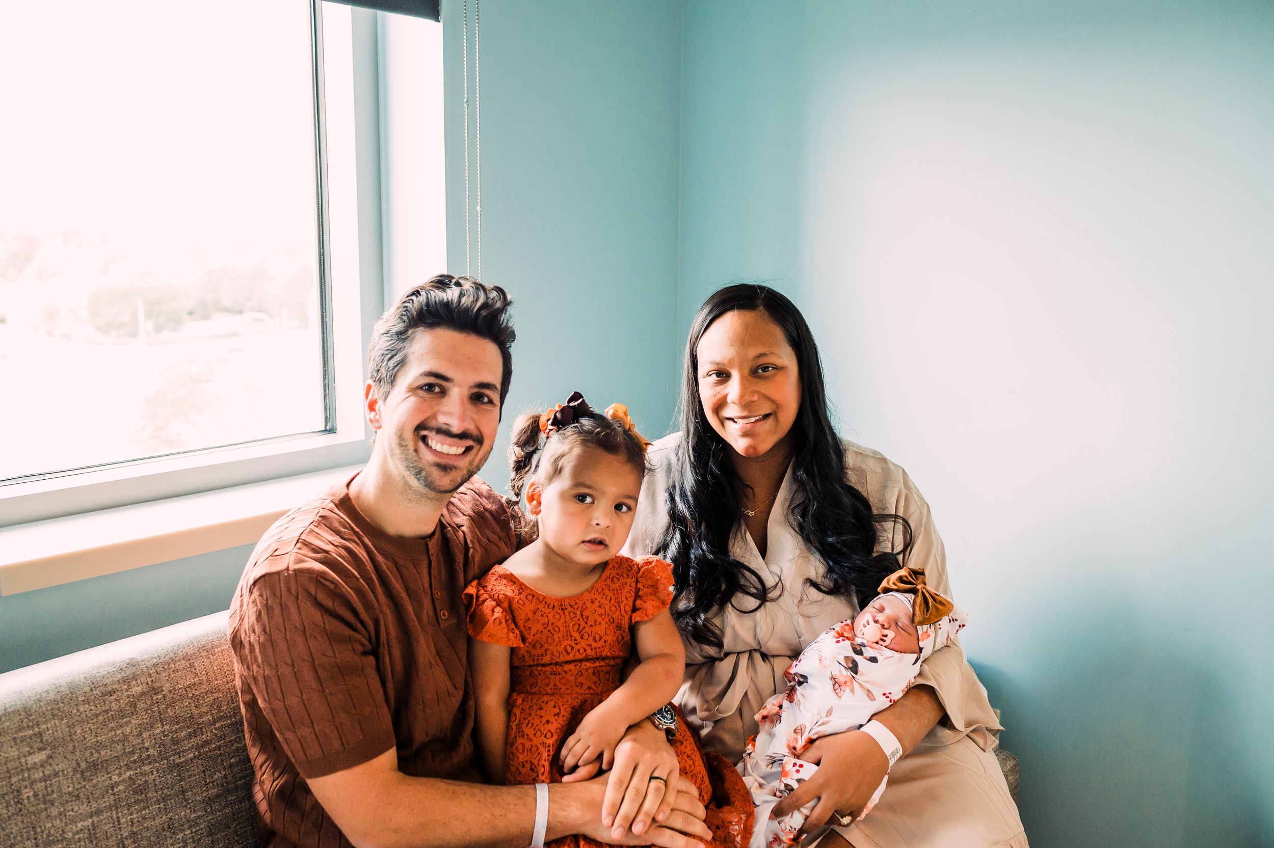 Family sitting together on gray couch near window in room with teal walls.