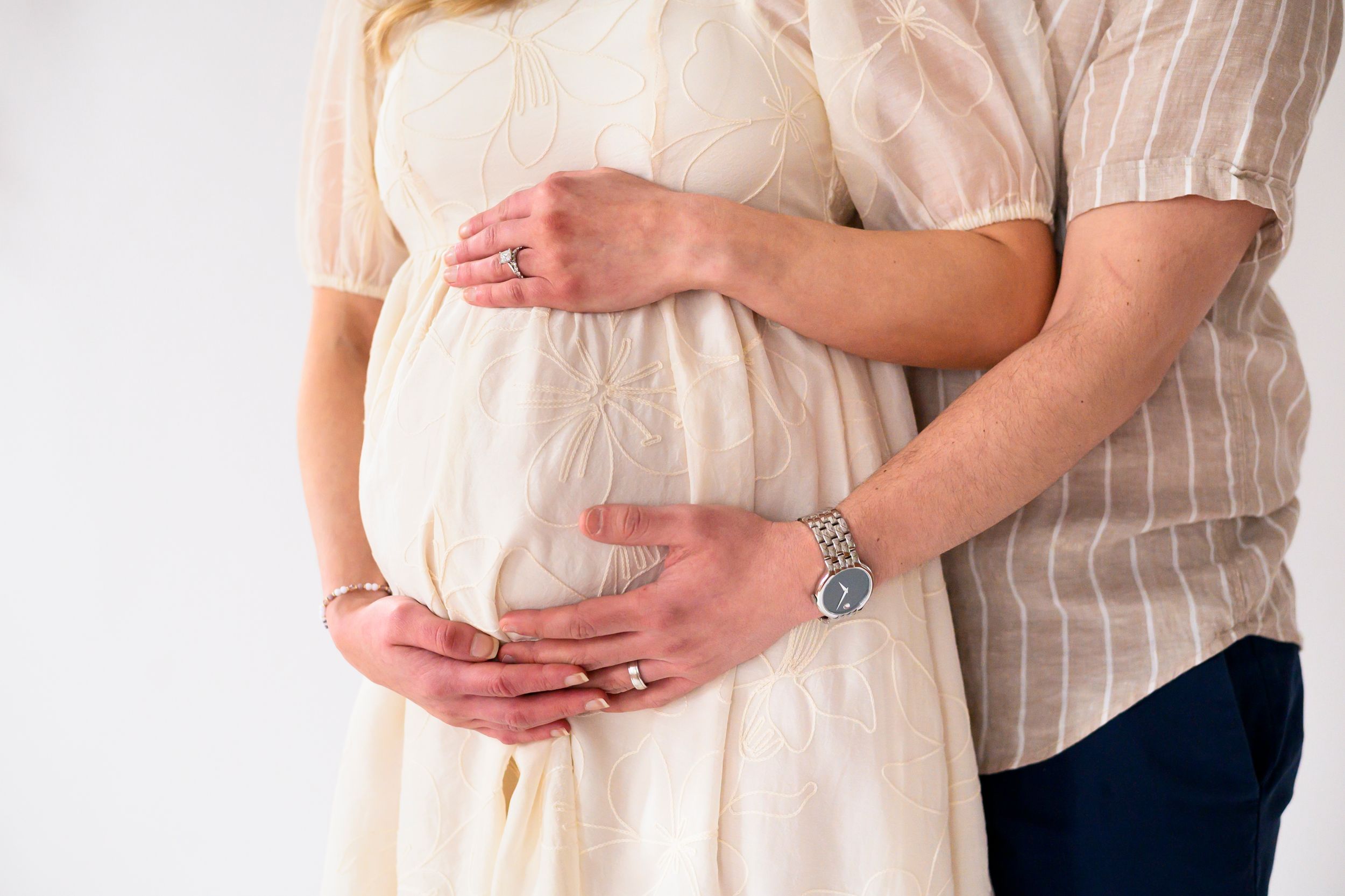 Close-up of pregnant belly being gently cradled by hands wearing a silver watch against white clothing.