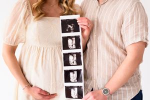 Couple in light colored outfits holds strip of ultrasound photos against pregnant belly in a maternity announcement photo.