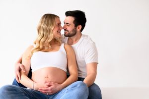 A couple sit close together on the floor against a white wall, with the woman wearing a white crop top showing her pregnant belly.
