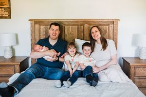A family sits together on a bed with wooden headboard, wearing coordinated white and navy blue outfits.