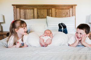Three children lie together on a bed with wooden headboard, showing playful expressions.