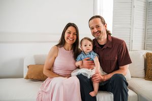 Three people sit together smiling on a white couch in a bright living room setting.