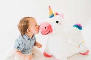 A child in denim plays with a large plush rainbow unicorn toy against a white background.