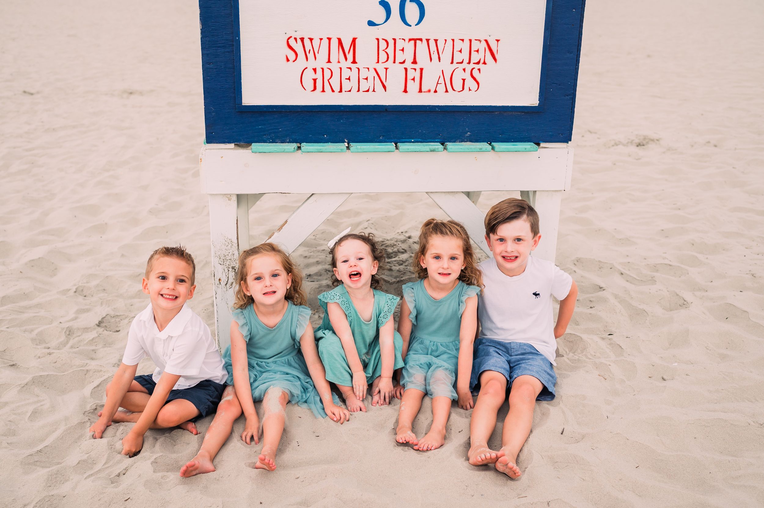 A group of five children in matching aqua and blue outfits sit together on a beach under a swim safety sign.