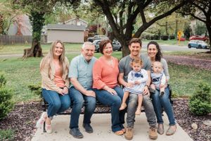A multi-generational family sits together on a wooden bench in a park setting during autumn.