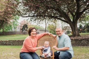 Happy family with toddler enjoying time together in wicker chair under blooming tree in garden setting.