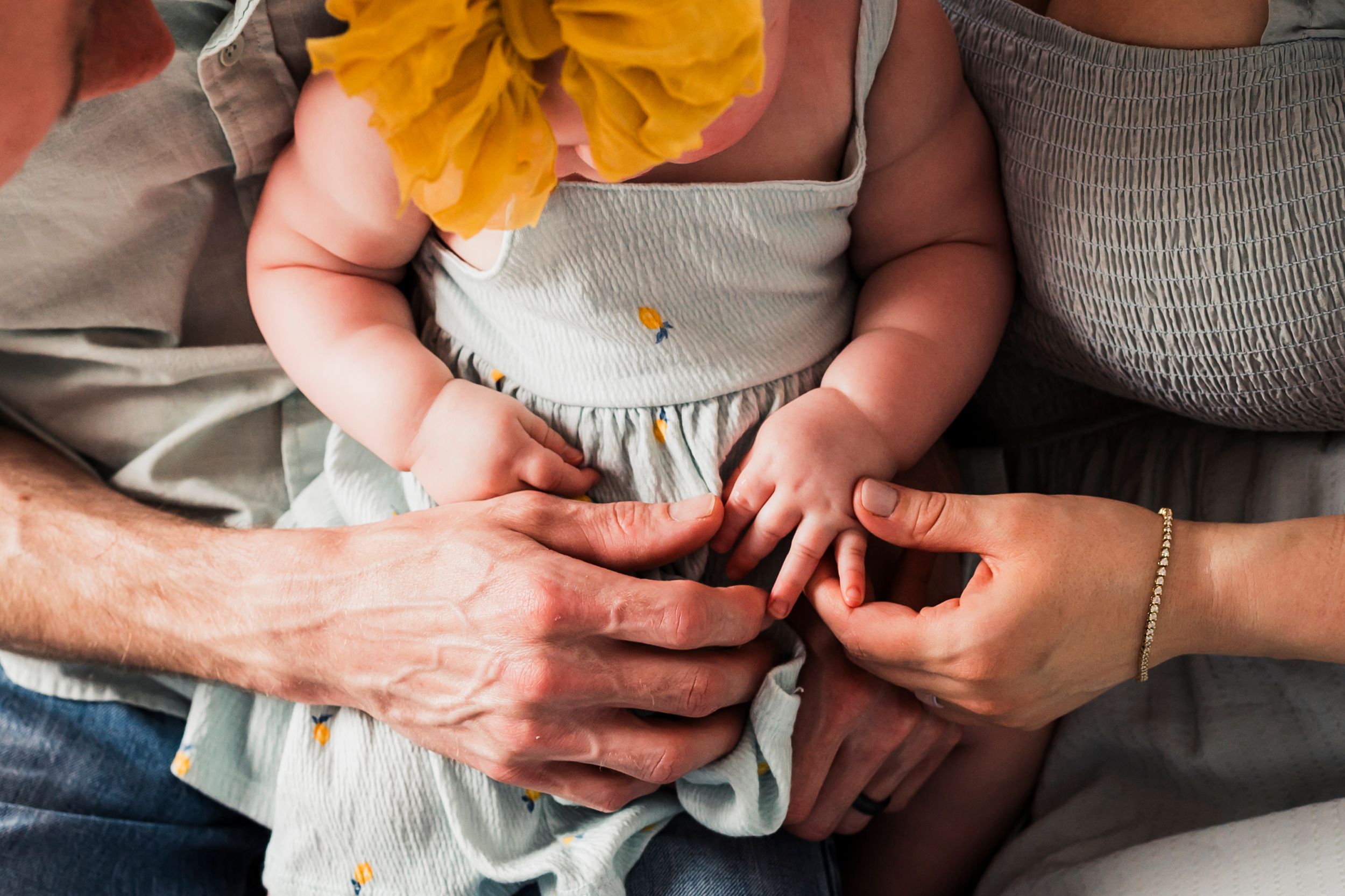 Hands gently hold a baby wearing a light gray dress with a yellow flower decoration.