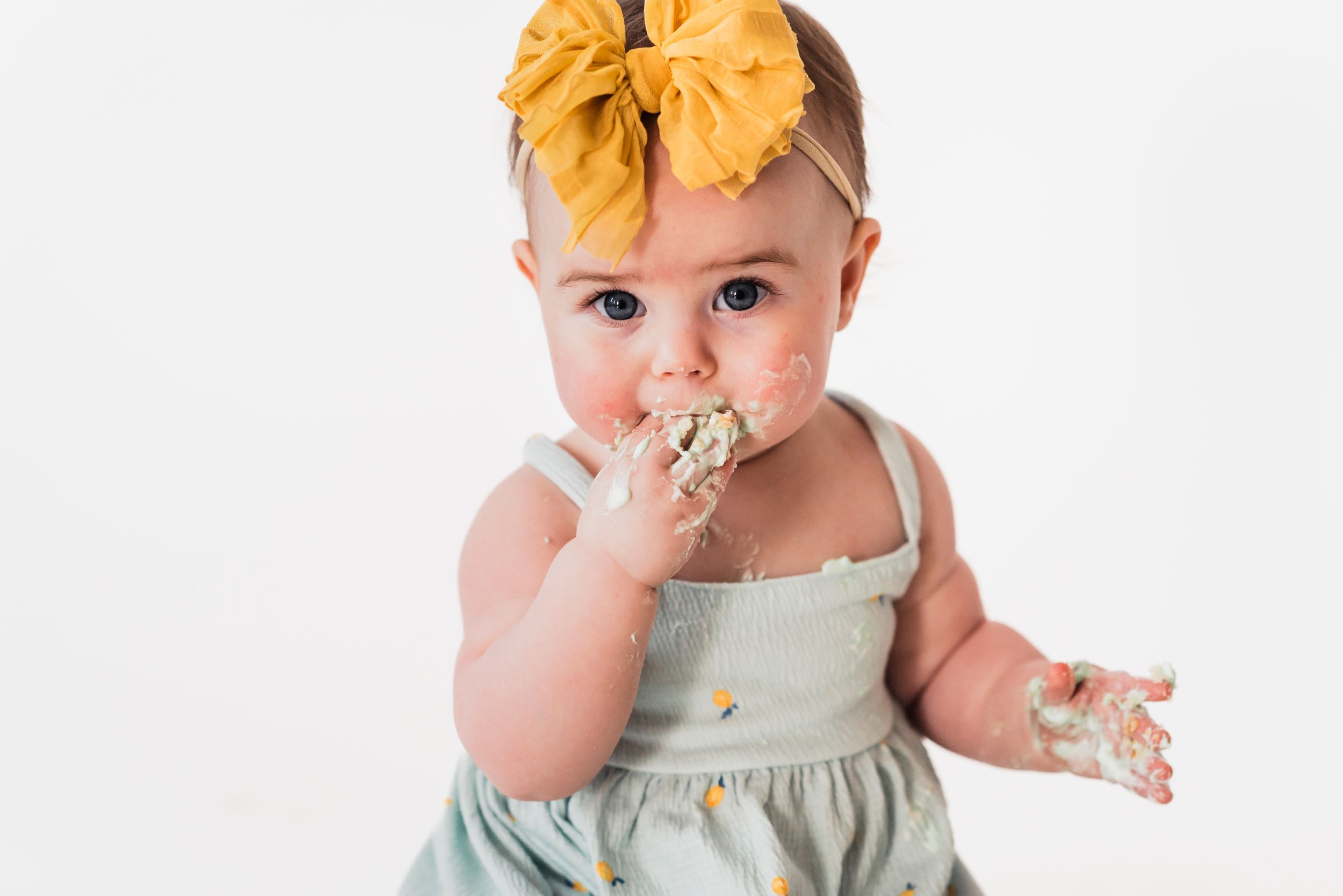 A baby wearing a mint green dress and yellow bow headband enjoys birthday cake with messy frosting-covered hands.