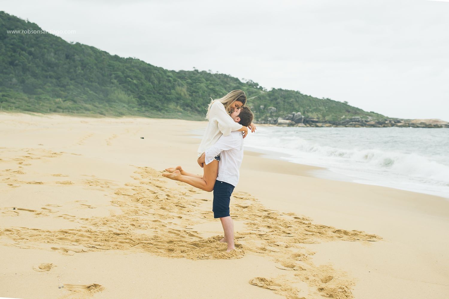 A couple embraces and dances together on a sandy beach with green hills and ocean waves in the background.