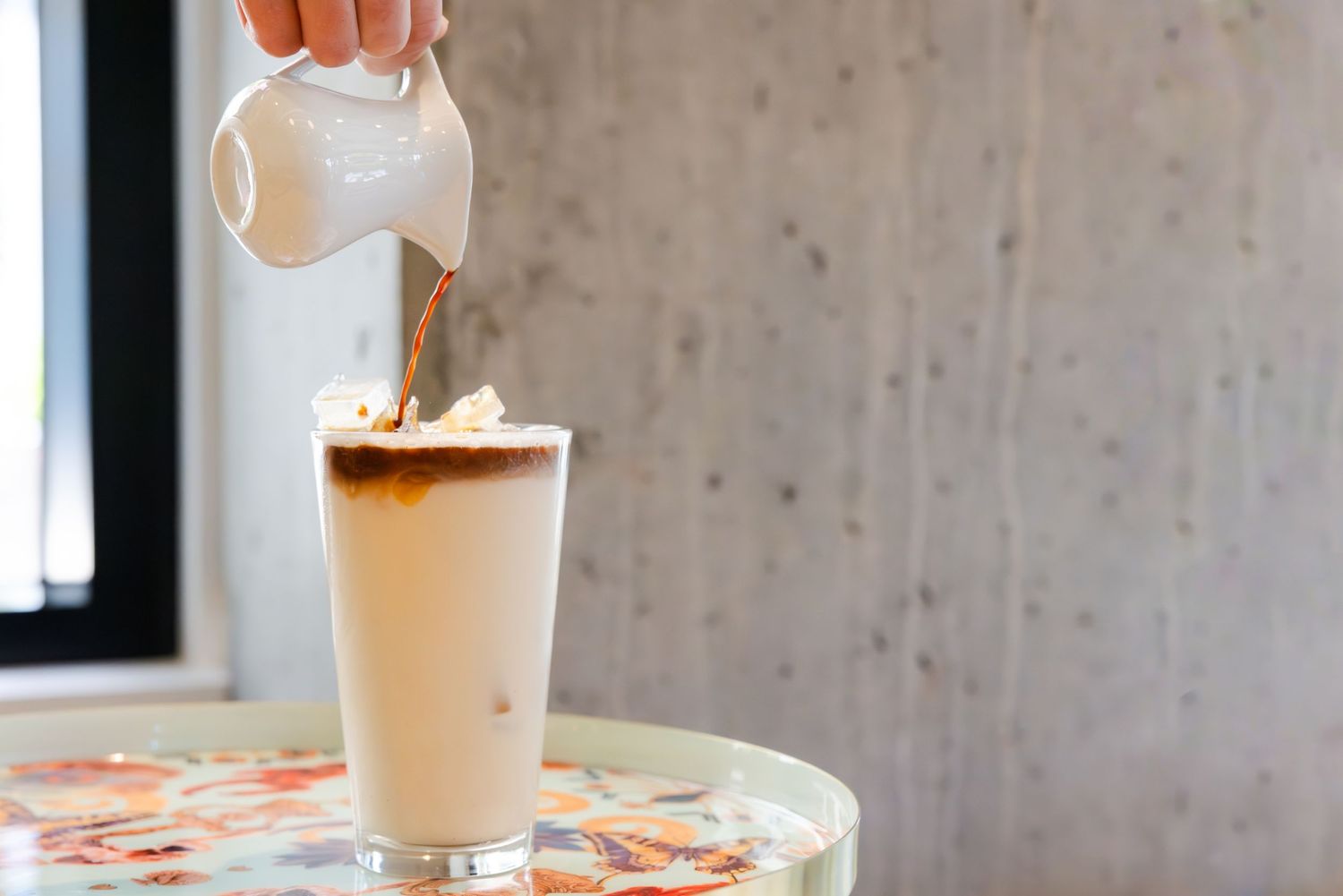 Cream being poured into a tall glass of iced coffee drink on a floral tablecloth.