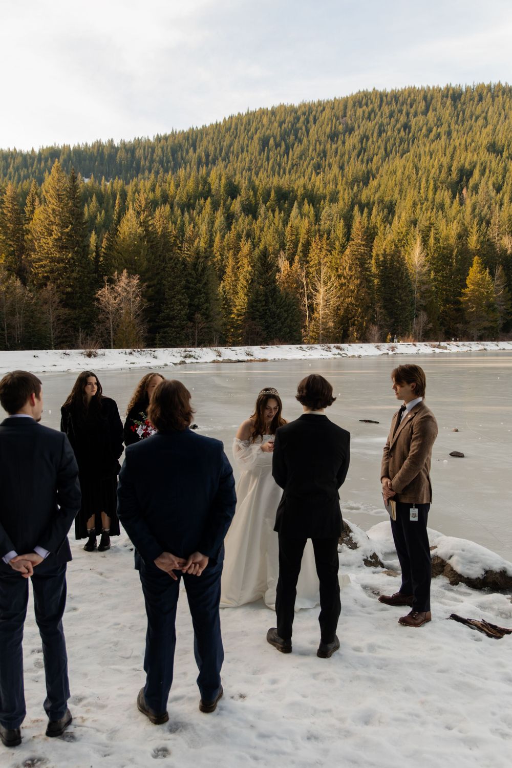 Winter wedding ceremony with guests gathered on snowy shore beside evergreen forest.