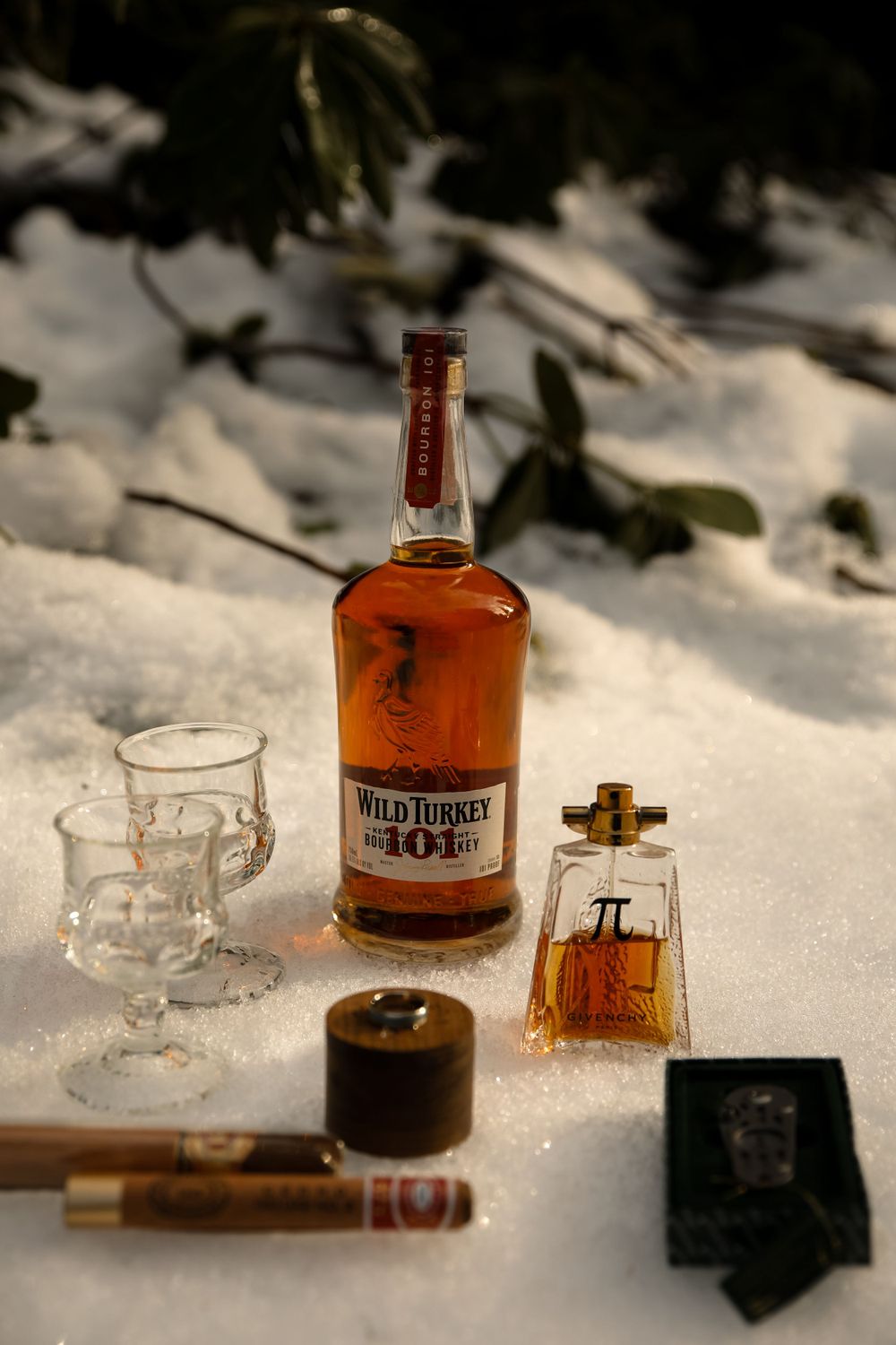 Wild Turkey bourbon bottle with glasses and cigar arranged in snow.