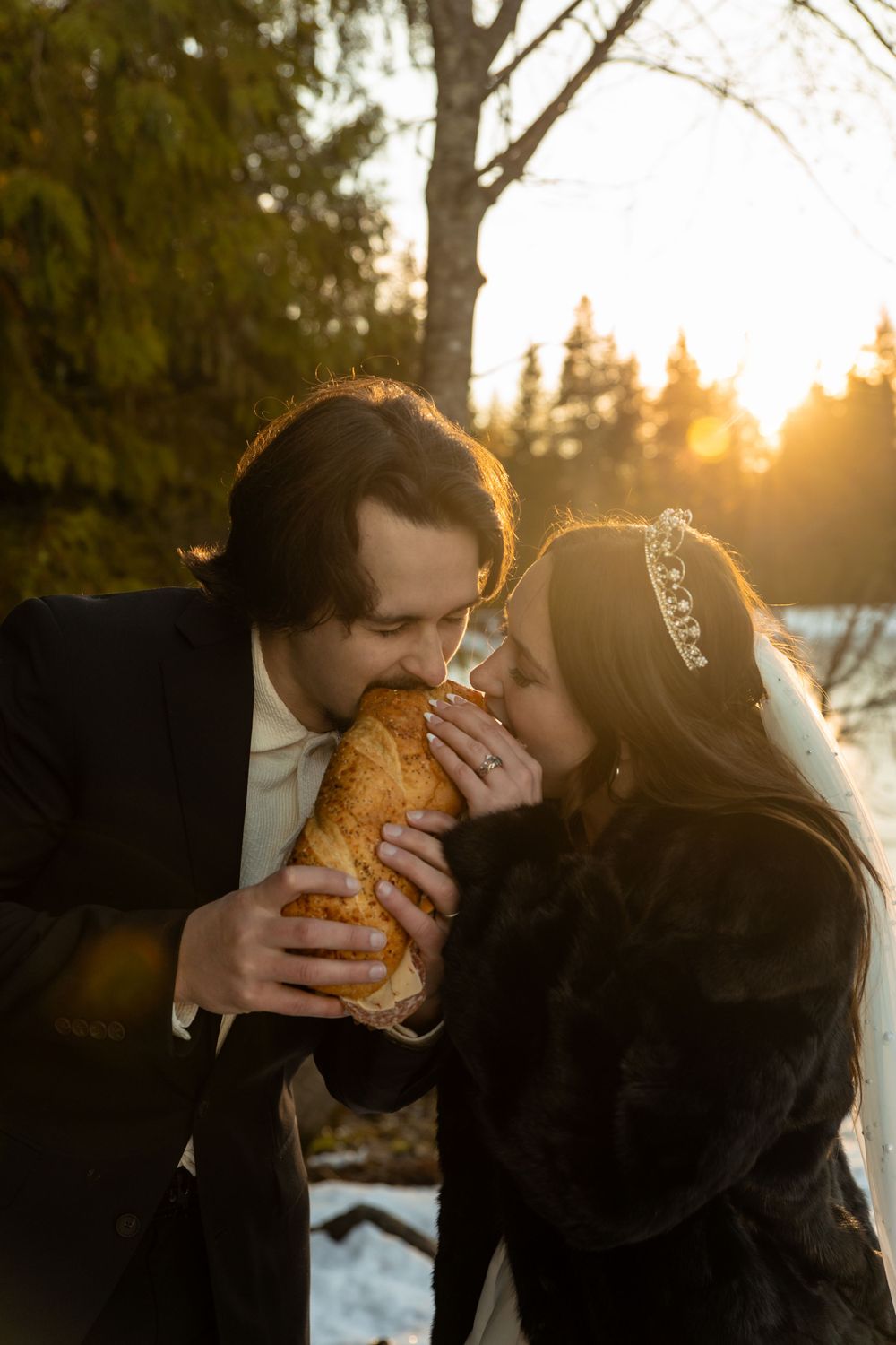 Couple shares a baguette together during golden hour in a winter forest setting.