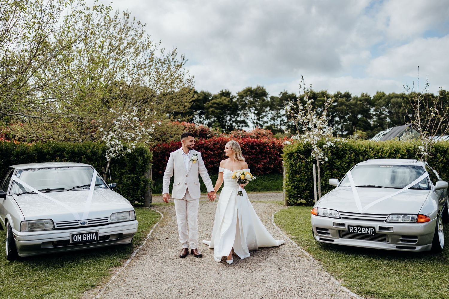 Bride and groom walking hand in hand between two classic cars decorated with white ribbons at an outdoor wedding venue.