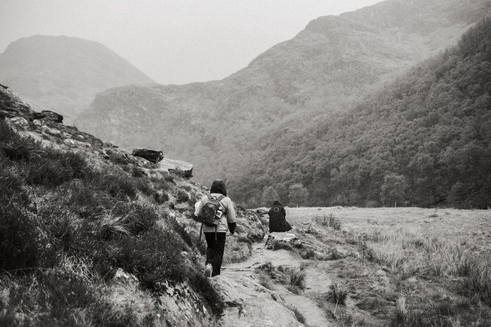 Two hikers trekking along a mountain trail surrounded by lush forested hills in a black and white landscape photo.