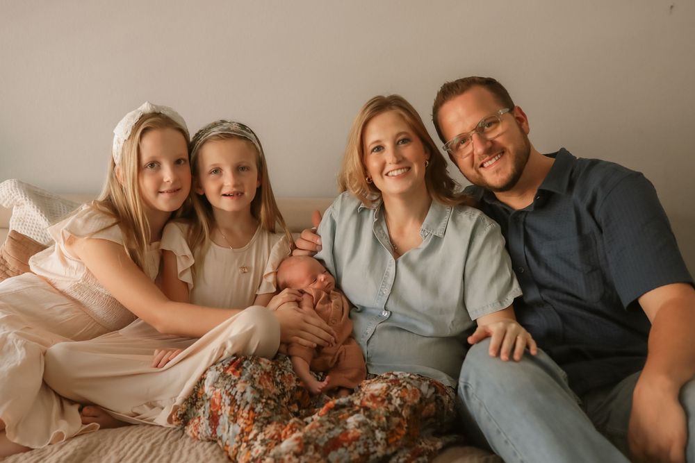 Happy family of five posing together, parents holding newborn baby, with two young daughters smiling warmly.