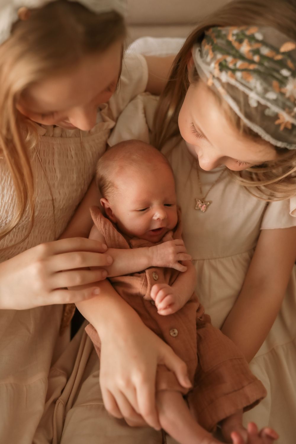 Two young girls lovingly holding a smiling newborn baby dressed in a brown outfit.