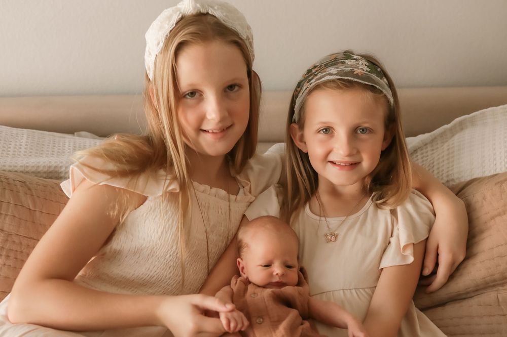 Two older sisters in cream dresses lovingly hold their newborn sibling during a soft, neutral-toned family photo session.