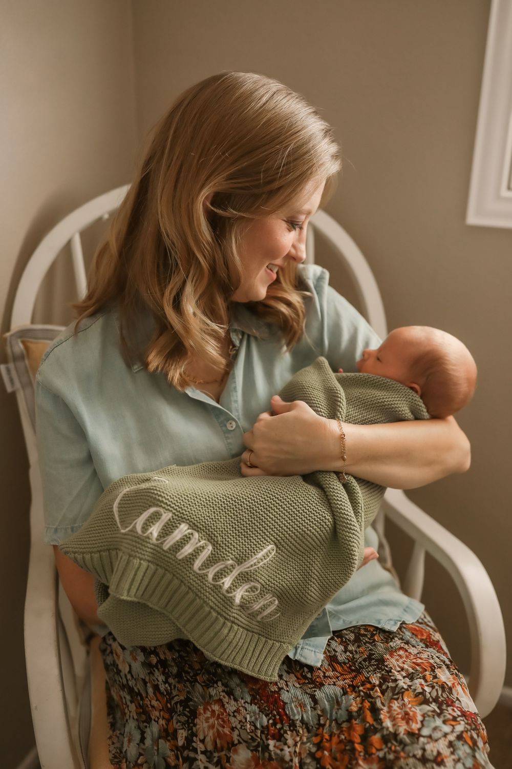 A smiling mother sits in a white rocking chair holding her newborn wrapped in a green knit blanket personalized with the name Camden.