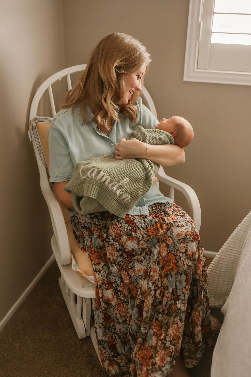 Mother sitting in white rocking chair holding newborn wrapped in sage green knit blanket embroidered with name Camden.