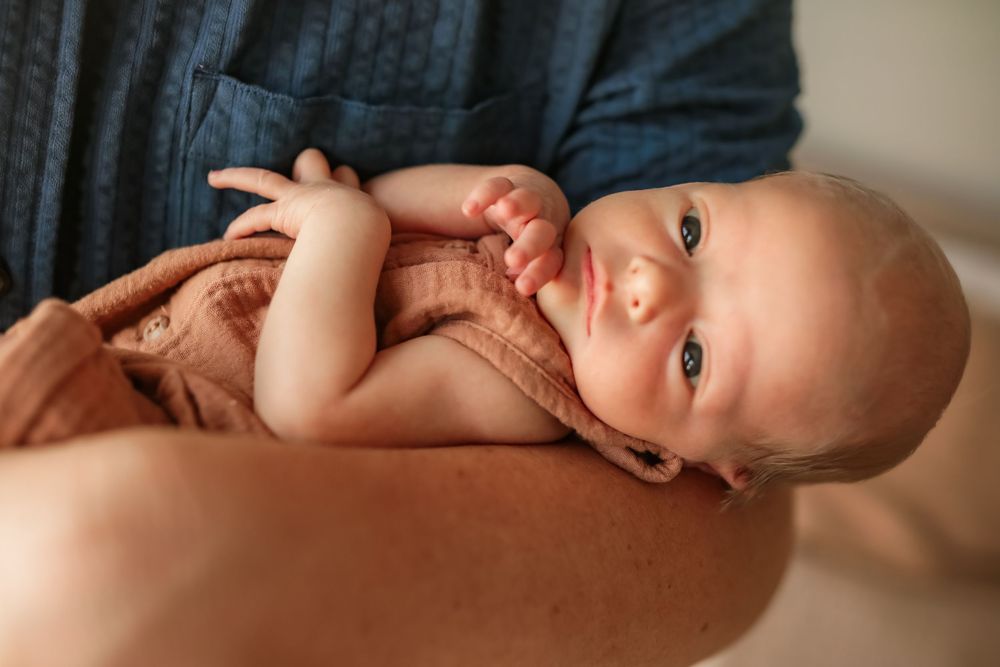 Newborn baby with blue eyes wrapped in a rust blanket cradled in an adult's arms.