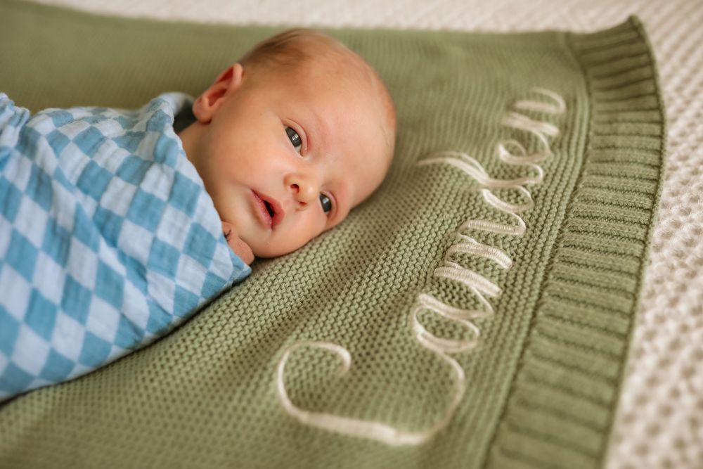 Newborn baby lying on a personalized green knit blanket named Camden, wrapped in a blue argyle swaddle.